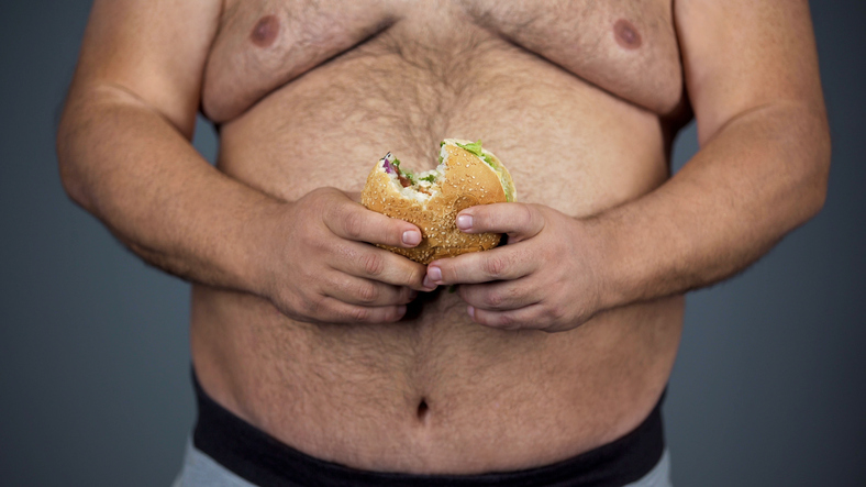 Niewłaściwa dieta jako przyczyna. Źródło zdjęcia: Getty Images Mężczyzna, duży brzuch i hamburger