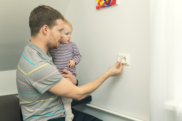 Używanie zatyczek bezpieczeństwa zmniejsza ryzyko obrażeń. Zdjęcie: Thinkstock Tata trzyma małe dziecko w ramionach i zamyka wtyczkę elektryczną