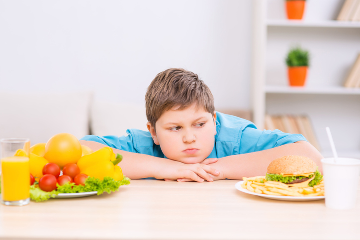 Żadne dziecko nie je warzyw, ponieważ są one zdrowe. Jedzą je, ponieważ ich rodzice również lubią je jeść. Zdjęcie: Thinkstock Gruby dzieciak między dwoma talerzami ze zdrowym i niezdrowym jedzeniem.