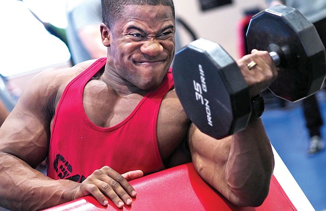Białka są dobrym suplementem diety do budowania masy mięśniowej. Źródło zdjęcia: Pixabay Mężczyzna w czerwonym podkoszulku ćwiczy biceps ze sztangą na jednym ramieniu.